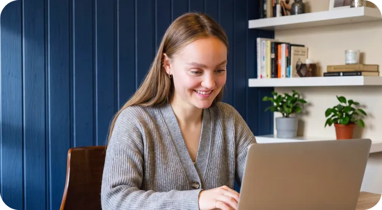 A young woman smiling as she uses her laptop to learn about what IBS is and everything she needs to know.