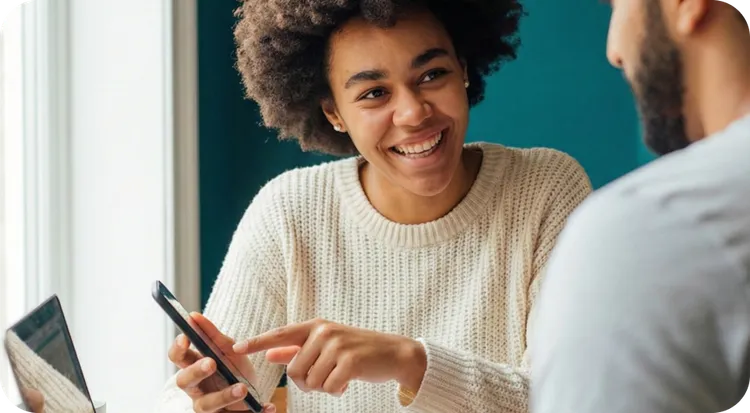 Mujer joven con pelo rizado y jersey blanco sonriendo ampliamente y señalando la pantalla de su smartphone mientras interactúa con un compañero o cliente