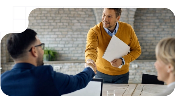 Man standing in a mustard sweater shaking hands with a man in a blue suit seated in front of a laptop after signing a confidentiality agreement.