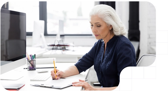 Older woman writing on a clipboard at her office desk.